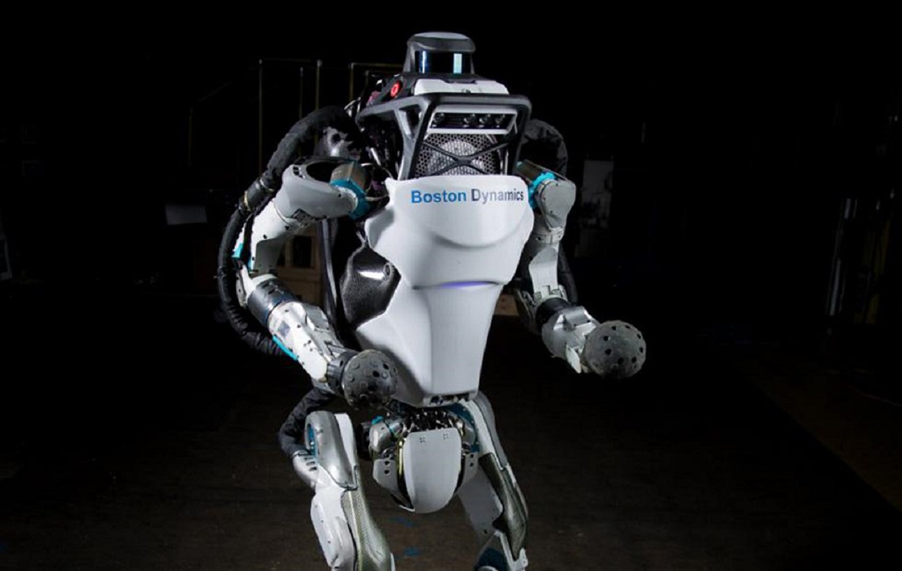  A humanoid robot with the name Boston Dynamics Atlas printed on its chest stands in a dark room.